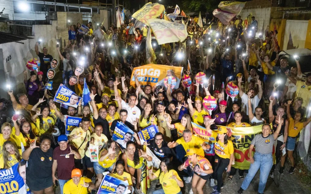Lucas Ribeiro cercado por grande multidão de apoiadores com luzes de celulares e bandeira laranja de Guga no Valentina.