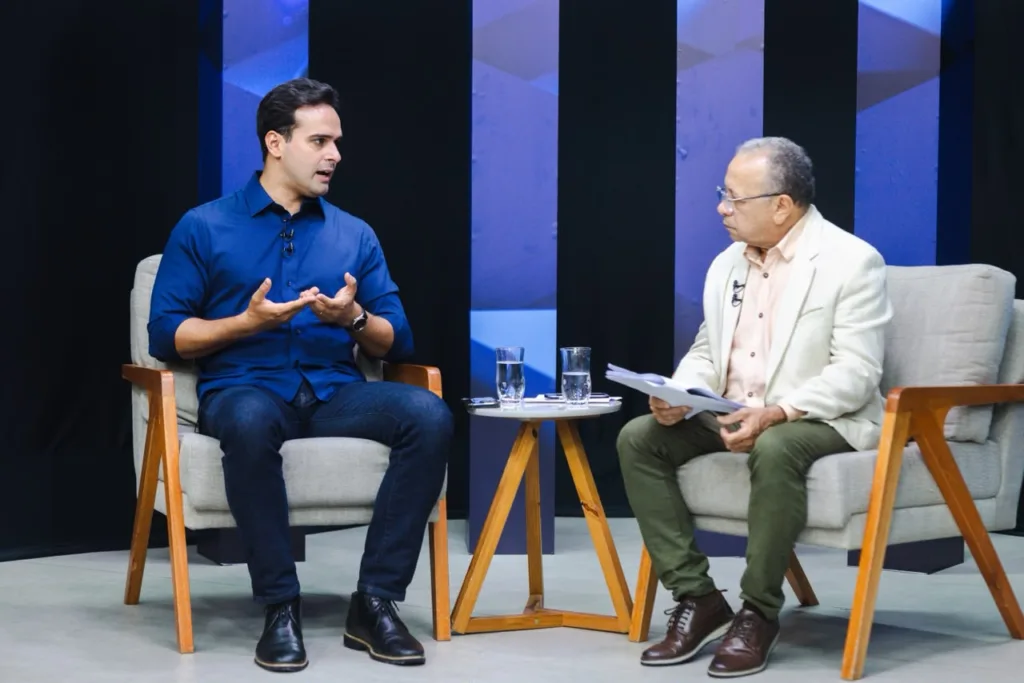 Lucas Ribeiro sentado em poltrona azul conversa com jornalista em estúdio de televisão durante programa de entrevistas.