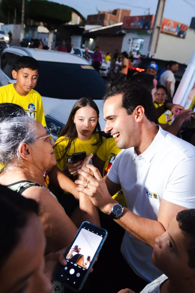 Lucas Ribeiro segura as mãos de uma idosa de óculos em momento afetuoso cercado por crianças e apoiadores.