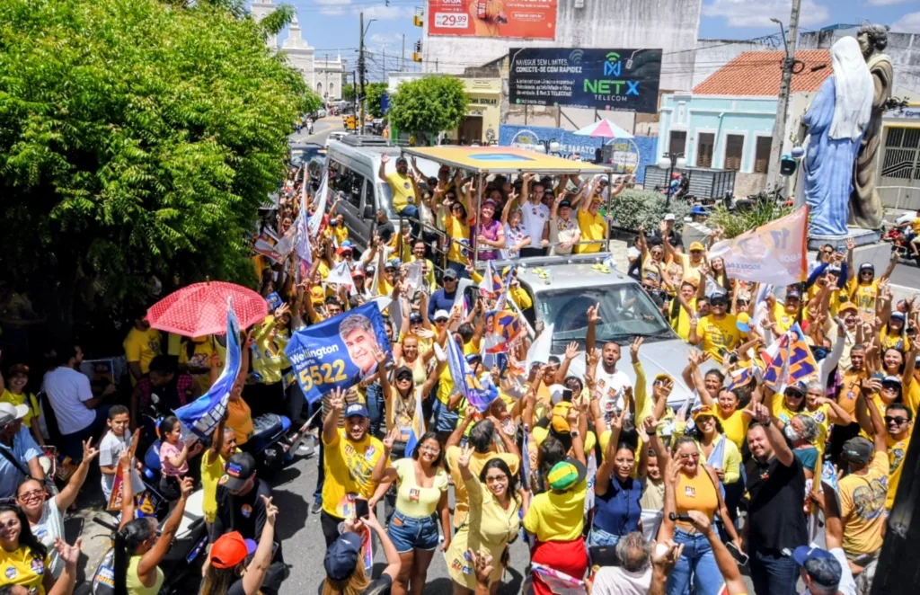 Vista aérea de carro aberto com a comitiva cercado por dezenas de apoiadores com bandeiras e sombrinha vermelha na rua.