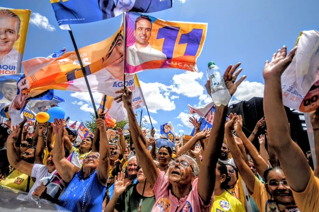 Multidão de apoiadores ergue as mãos com bandeiras de campanha sob céu azul em Uiraúna.