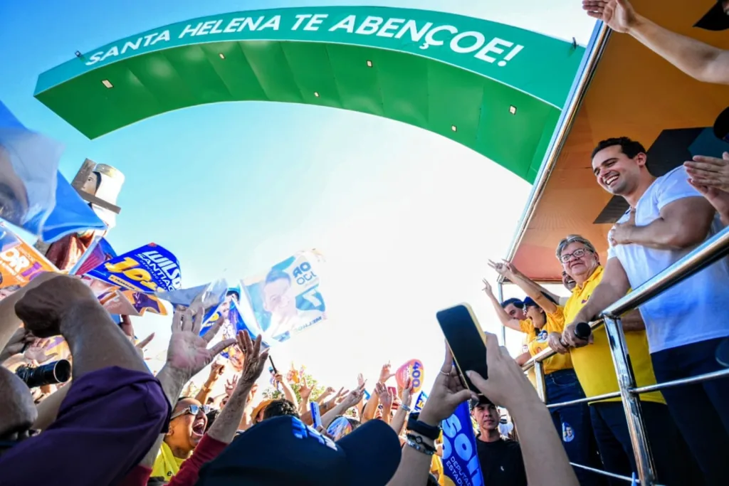 Lucas Ribeiro com a mão no peito e João Azevêdo sorrindo em carro aberto sob portal verde de Santa Helena diante da multidão.