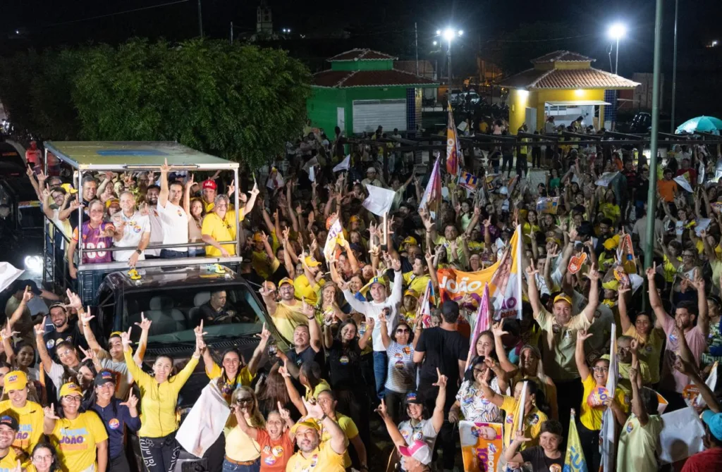 Imagem aérea noturna mostrando dezenas de apoiadores com as mãos levantadas cercando veículo com Lucas Ribeiro e lideranças.