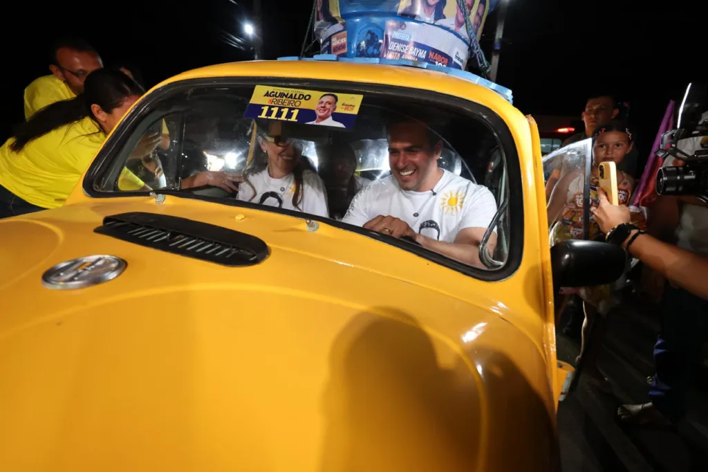 Lucas Ribeiro sorri dirigindo fusca amarelo ao lado da prefeita Denise Bayma durante mobilização política à noite.