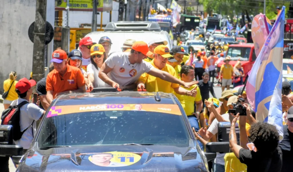 Lucas Ribeiro estica o braço para cumprimentar apoiadores ao lado de Nabor Wanderley e João Azevêdo em carro aberto.