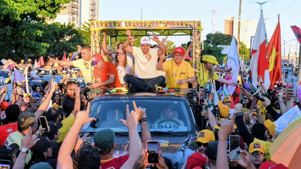 Lucas Ribeiro sentado no teto do carro faz sinal do 'L' com os dedos ao lado de João Azevêdo e Nabor Wanderley.